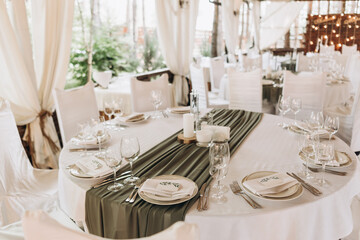 Fototapeta na wymiar Wedding table setting. Banquet table with white tablecloths served with white plates, silver cutlery, white napkins and wine glasses. olive colore drapery on a white set table