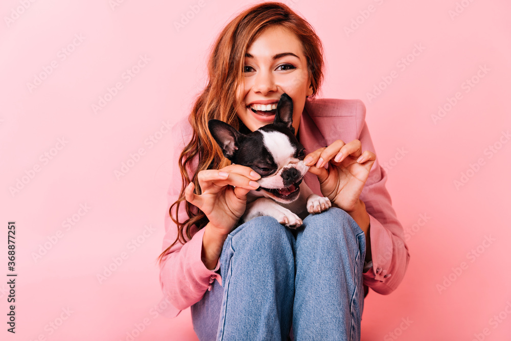 Wall mural Glamorous woman touching her dog's cheek. Indoor photo of blissful female model sitting on pink background with puppy. - Wall murals