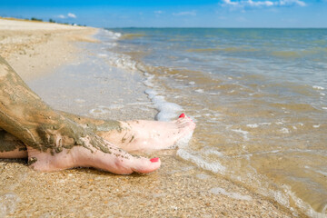 View of beautiful female feet with bright red pedicure on the sand of the beach. The ankles of the feet are smeared with healing mud. Skin rejuvenation while relaxing at sea. Spa. Copy space.