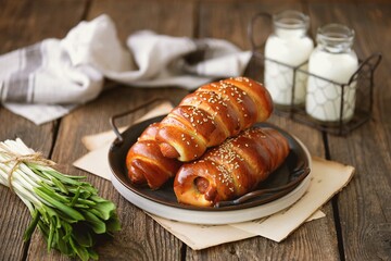 Homemade sausages in yeast dough with sesame seeds and milk on a wooden background.