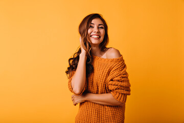 Adorable woman in orange attire touching her brown wavy hair. Laughing blithesome girl posing on yellow background. - obrazy, fototapety, plakaty