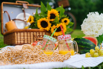 Summer picnic concept on sunny day with watermelon, fruit, bouquet hydrangea and sunflowers flowers. Picnic basket on grass with food and refreshing summer drink on white knit blanket. Selective focus