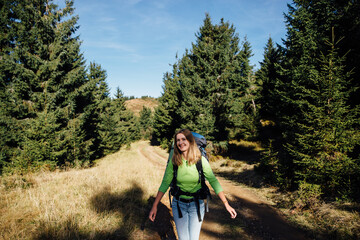 Happy female hiker walking with big heavy backpack in autumn mountains forest looking at camera and smiling