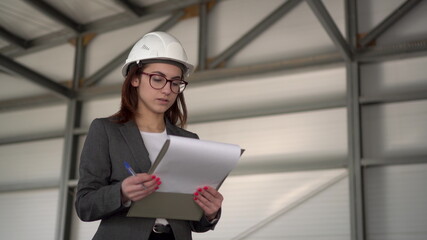 Young woman in a helmet with documents at a construction site. The boss woman in a suit keeps records of documents on architecture. The girl looks around.