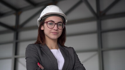 A young woman in a protective helmet crossed her arms and stands at a construction site. The boss woman in a suit looks at the camera.