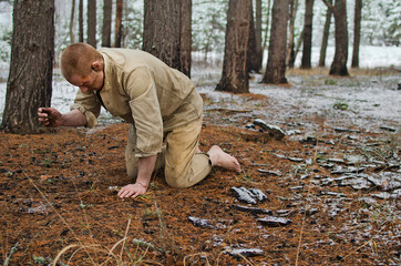 young man in a world war II uniform and barefoot kneels in a winter forest