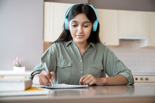 Young indian teen girl wearing headphones writing homework in notebook studying at home listening audio course remote learning making notes working or distant learning sitting at table at home office. - Powered by Adobe