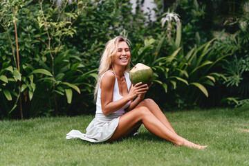 Beautiful blonde woman in the white dress on the tropical terrace with coconut drink in the rays of...