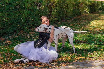 Fototapeta premium Ballerina with Dalmatian dog in the Park. Woman ballerina in a white ballet skirt and black leather jacket and in pointe shoes in autumn park hugging her spotty dalmatian dog.