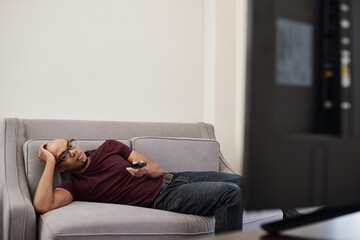 Obraz premium Tired or bored young Black man lying on sofa and changing tv channels when trying to find what to watch