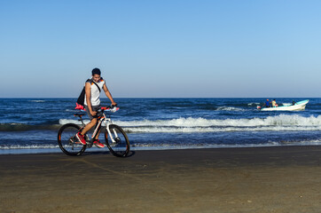 Fototapeta na wymiar joven montado en bici en la playa sonriendo y una lancha de fondo