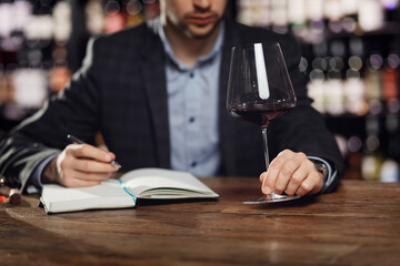 Sommelier compiles tasting card of alcohol for restaurant, evaluates red wine in glass