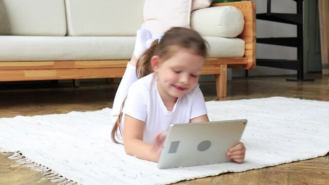 Child girl using digital tablet lying on carpet floor alone, holding and surfing touchpad internet at home.