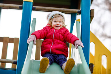 Fototapeta na wymiar Cute toddler girl having fun on playground. Happy healthy little child climbing, swinging and sliding on different equipment. On cold day in colorful clothes. Active outdoors game for children