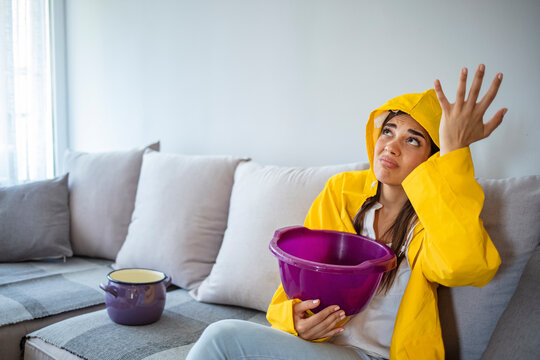 Worried young woman in raincoat holding bucket under water drops. Upset young woman using bucket during leak in living room. Frustrated brunette woman sitting on sofa under leaking ceiling