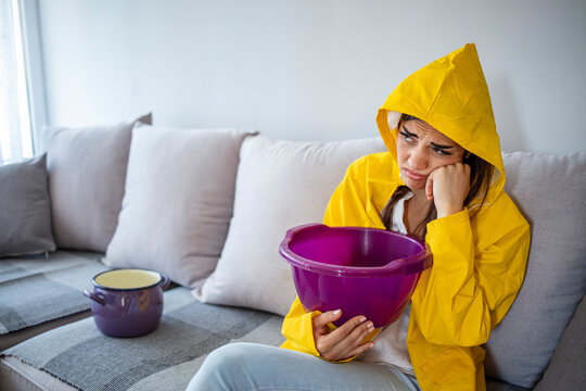 Woman in raincoat waiting for plumber to fix flood. Frustrated female client in raincoat holding pot while water leaking from roof in hotel. Woman collecting leaking water from ceiling in living room