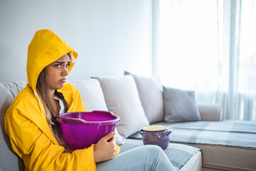 Upset woman touching head while sitting on sofa in raincoat under leaking ceiling. Upset woman...