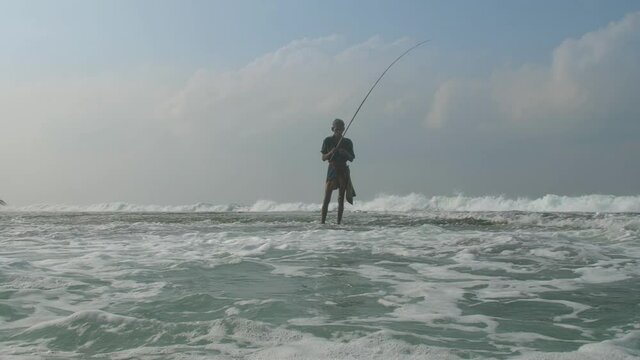 aged Sinhalese fisherman stands in foaming ocean water near beach under cloudy sky in summer slow motion. Concept aboriginal people