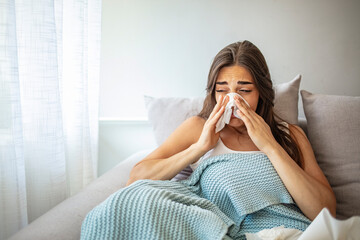 Woman Suffering From Cold Lying In Bed With Tissue. Sick young woman at home on the sofa with a cold, she is covering with a blanket and blowing her nose