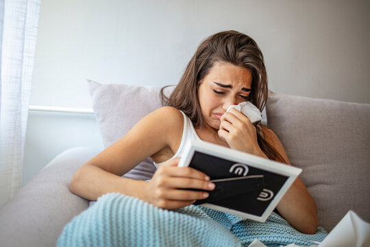 Sad woman sitting on grey couch, looking at photo frame and crying at home, grieving disorder concept. Young woman sitting crying and see photo in the frame on the couch, copy space