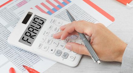 Fraud text on a calculator with financial data paperwork on the desk. Business and finance fraud prevention or identification concept. Hand with calculator