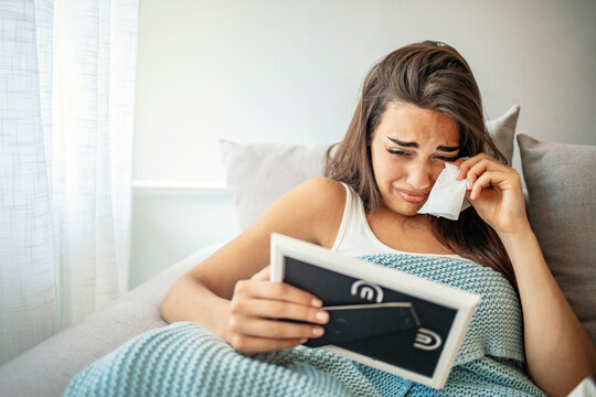 Upset woman lying in bed, looking at picture frame, crying and wiping tears at home. Depressed young woman suffering from break up with boyfriend or divorce with husband