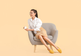 Young woman with coffee relaxing in armchair on color background