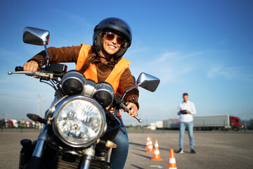 Female student with helmet taking motorcycle lessons and practicing ride. In background traffic cones and instructor with checklist rating and evaluating the ride. Motorcycle school of driving. - obrazy, fototapety, plakaty