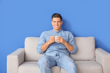 Young man with tea relaxing on sofa near color wall