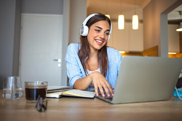Smiling girl student wear wireless headphone study online with skype teacher, happy young woman learn language listen lecture watch webinar write notes look at laptop sit in cafe, distant education