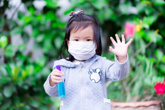 Asian girl wearing white face mask, in her hand holding a bottle of blue alcohol and lifting her hand, forbidding other people to come close to herself in measure with Social Distancing in Covid-19.