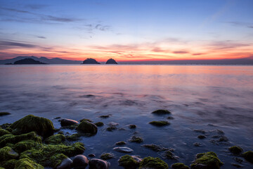 Naklejka premium Amazing seashore sunrising,endless horising and green mossy round gravel pebble beach, sea waves beats on a rocky shore with foam.
