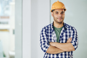Portrait of handsome smiling contractor in hardhat folding arms and looking at camera