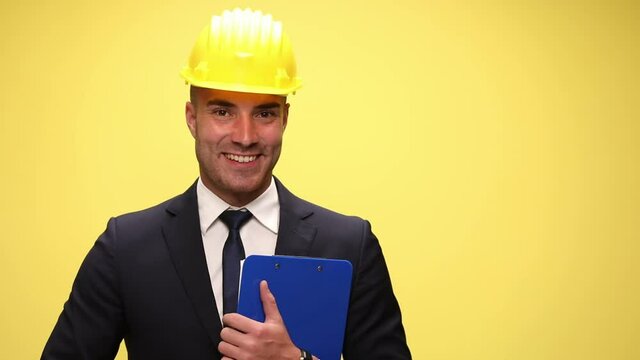smiling elegant engineer in suit wearing safety helmet, reading from clipboard and presenting, holding hands in pockets and adjusting suit on yellow background