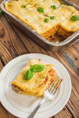 Homemade lasagna ready to eat in metal baking tray served with basil leaves, tomatoes, and turquoise tablecloth on brown wooden background. One portion on the white plate with fork. Top view.