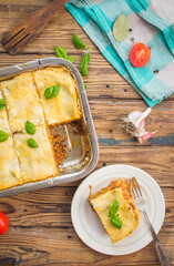 Homemade lasagna ready to eat in metal baking tray served with basil leaves, tomatoes, and turquoise tablecloth on brown wooden background. One portion on the white plate with fork. Top view.