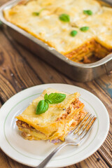 Homemade lasagna ready to eat in metal baking tray served with basil leaves, tomatoes, and turquoise tablecloth on brown wooden background. One portion on the white plate with fork. Top view.