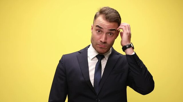 clueless young businessman in suit looking to side, scratching head and thinking, having an idea and pointing finger, smiling and standing on yellow background