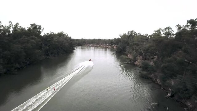 Aerial reveal shot of speed boat with 2 skiers following boat at a very fast pace upstream of a river on a cloudy day