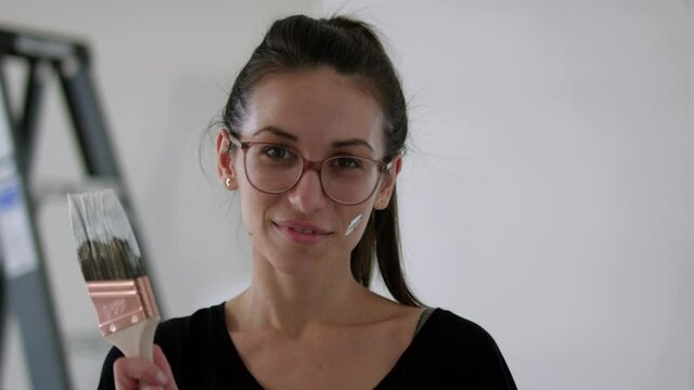 Woman holding paintbrush in hand painting room - looking right into camera - medium shot