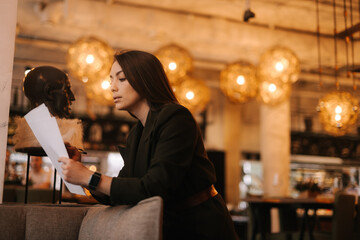 Professional young woman inspector wearing stylish formal clothes in modern luxury restaurant writing in paper documents with pen. Business lady lady in suit posing in cafe.