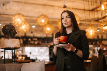 Beautiful young woman holding cup of tasty hot coffee in hand while standing in restaurant. Caucasian lady posing with glass of beverage in cafe with modern dark interior. Shotting from below.