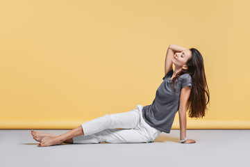 Barefoot young woman in casual trousers and shirt sits on the floor in studio on yellow background