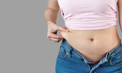 Naklejka premium Woman in unbuttoned jeans holds with hand and squeezes excess belly fat. The concept of overweight, weight loss, diet, unhealthy lifestyle, obesity, junk food on gray background, banner, copy space.