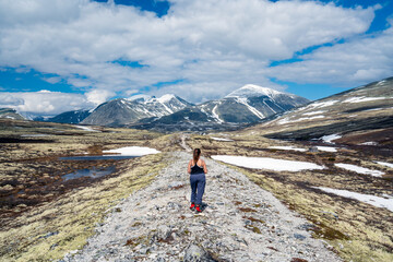 Female hiker stands on gravel path overlooking beautiful mountain scenery with snow capped peaks and blue sky.