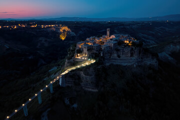 Fototapeta premium Civita di Bagnoregio. Aerial, night view of ancient Italian city standing on top of a plateau, illuminated by lamps. Famously known as dying city. City on steep rock over Tiber river valley, Italy.