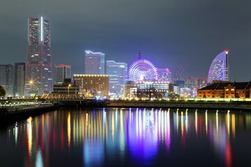 Fototapeta na wymiar Beautiful night scenery of Yokohama Minatomirai Bay area with high rise buildings in the background, a giant Ferris wheel in Cosmo World Amusement Park & colorful reflections of city lights on water