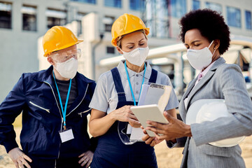 Black engineer and two workers with face masks cooperating while using digital tablet at construction site.