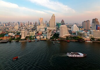 Aerial panorama of downtown Bangkok, vibrant capital city of Thailand, with a beautiful skyline of modern skyscrapers and busy traffic of ferries & tour boats on Chao Phraya River under sunset sky