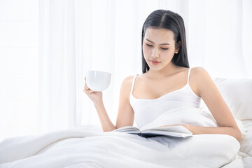 Obraz na płótnie Canvas A Asian sexy girl sitting on white bed in bedroom and drinking a cup of hot coffee, happy vacation morning